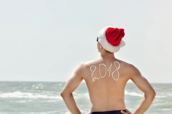 Mann mit Weihnachtsmütze mit der Aufschrift 2018 auf dem Rücken am Strand stehend — Stockfoto