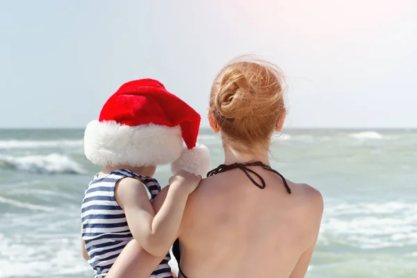 Kind mit Weihnachtsmütze in den Armen der Mutter, der Strand. zurück — Stockfoto