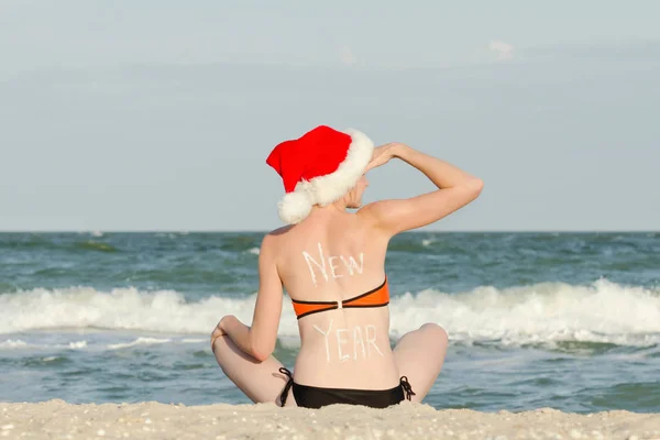 Yeni yıl geri üzerinde yazıt ile Noel Baba şapkalı kız sahilde oturuyor ve mesafe içine görünüyor — Stok fotoğraf