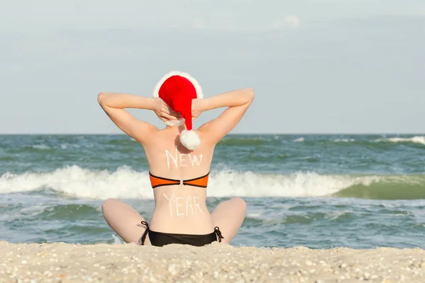 Mädchen mit Weihnachtsmütze mit der Aufschrift Neujahr auf dem Rücken am Strand sitzend — Stockfoto