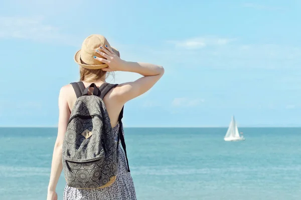 Una chica con un sombrero y una mochila en la costa. Velero en la distancia. Vista desde atrás — Foto de Stock
