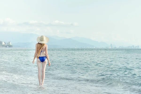 Menina em um maiô e chapéu caminha pelo mar. Vista de trás — Fotografia de Stock