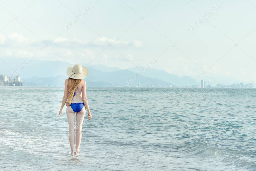 Girl in a bathing suit and hat walks by sea. View from the back