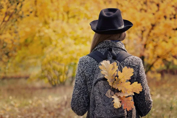 Menina em um chapéu com folhas amarelas em uma mochila está de pé na floresta de outono. Visão traseira — Fotografia de Stock