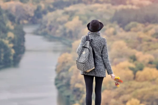 Chica con una mochila y un sombrero de pie en una colina. Río y bosque abajo. Vista trasera — Foto de Stock