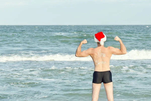 Starker Mann in Weihnachtsmannmützen mit der Aufschrift Neujahr auf dem Rücken, der am Strand steht. zurück — Stockfoto