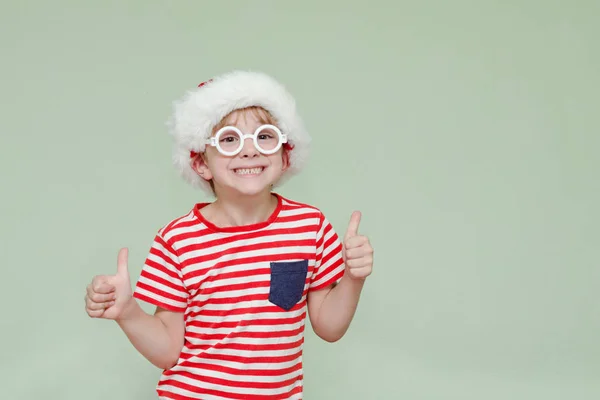 Chłopiec w Santa kapelusz i okulary, uśmiechając się i pokazuje kciuk w. — Zdjęcie stockowe