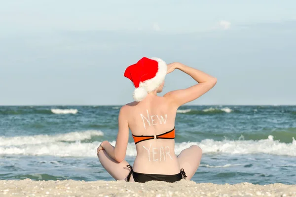 Mädchen mit Weihnachtsmütze mit der Aufschrift Neujahr auf dem Rücken am Strand sitzend — Stockfoto