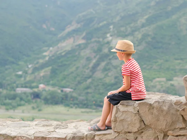 Boy in a hat sitting on stones on a background of green mountain — Stock Photo, Image