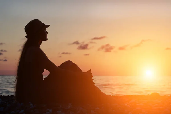 Девушка в шляпе, сидящая на фоне моря на закате — стоковое фото
