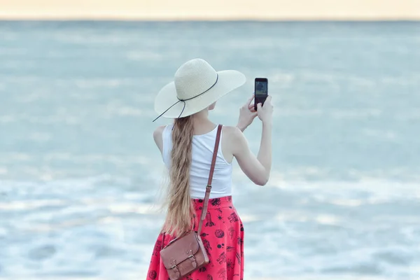 Девушка в красной юбке и шляпе делает селфи на пляже на фоне моря и неба. Вид сзади — стоковое фото