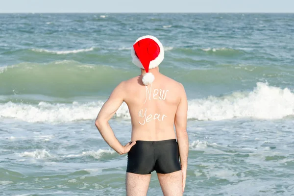 Человек в шляпе Санты с надписью Новый год на спине, стоящий на пляже — стоковое фото