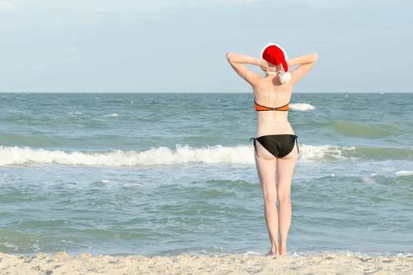 Menina na costa do mar com uma inscrição nas costas e chapéu de Papai Noel — Fotografia de Stock