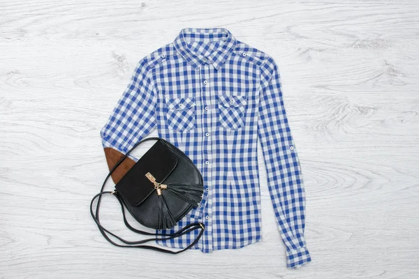 Μπλε και λευκό καρό πουκάμισο και μαύρη τσάντα. Ξύλινο υπόβαθρο. — Φωτογραφία Αρχείου