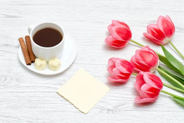 Pink tulips, mug of coffee and cinamon, light wooden background.