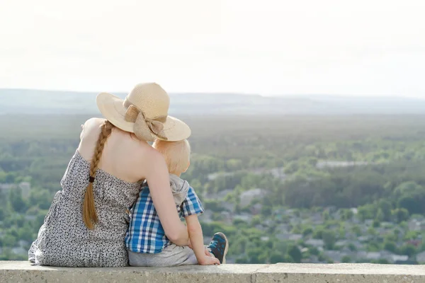 Anne ve oğlu yeşil orman ve köy uzakta arka plan üzerinde oturuyor. Arkadan Görünüm