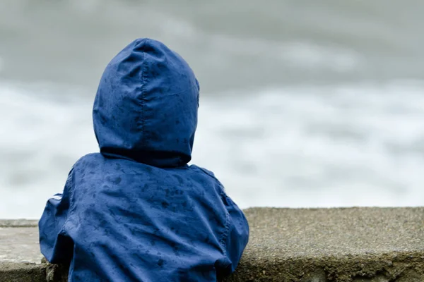 Kukuleta ile mavi ceketli çocuk sırtını duruyor. Pier deniz dalgaları arka planı — Stok fotoğraf