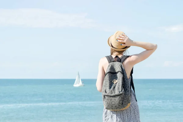Una chica con un sombrero y una mochila en la costa. Velero en la distancia. Vista trasera — Foto de Stock
