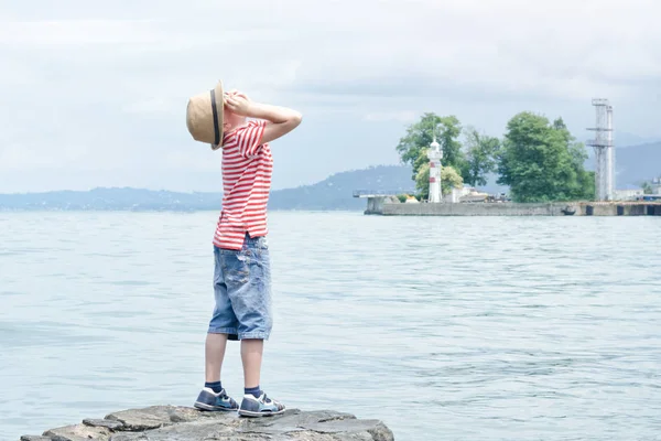Sahilde duran ve ararken şapkalı çocuk. Küçük deniz feneri — Stok fotoğraf
