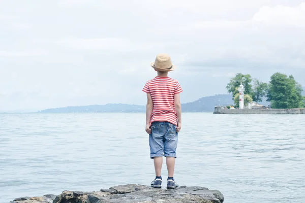 Çocuk şapka ayakta sahilde. Uzaktan küçük deniz feneri — Stok fotoğraf