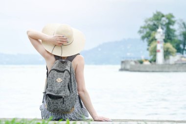 İskelede oturup bir sırt çantası ile şapkalı kız. Dağlar