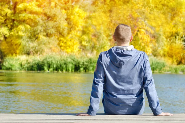 Young man is sitting on the dock. Autumn, sunny. Back view — Stock Photo, Image