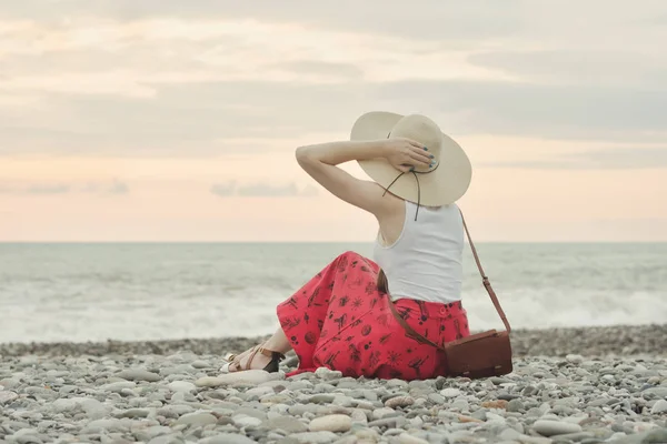 A menina de chapéu senta-se em uma praia de seixos. Vista traseira. Hora do pôr do sol — Fotografia de Stock