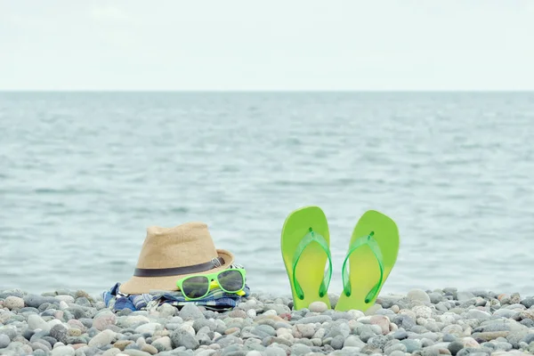 Шляпа, очки и шлепанцы на галечном пляже. Море на заднем плане — стоковое фото