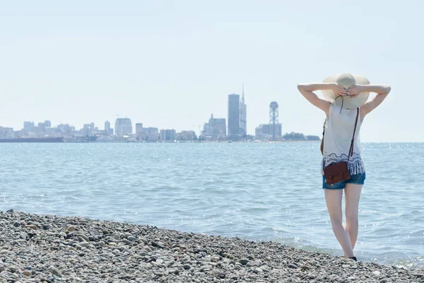 Una chica con sombrero a lo largo de la orilla del mar. Ciudad en la distancia — Foto de Stock