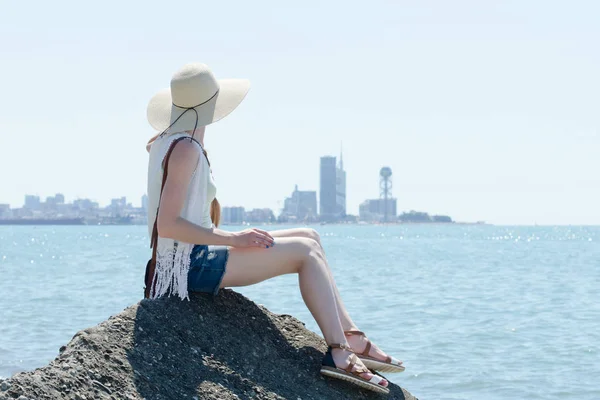 Chica con sombrero sentada en una roca junto al mar. Ciudad en la distancia — Foto de Stock