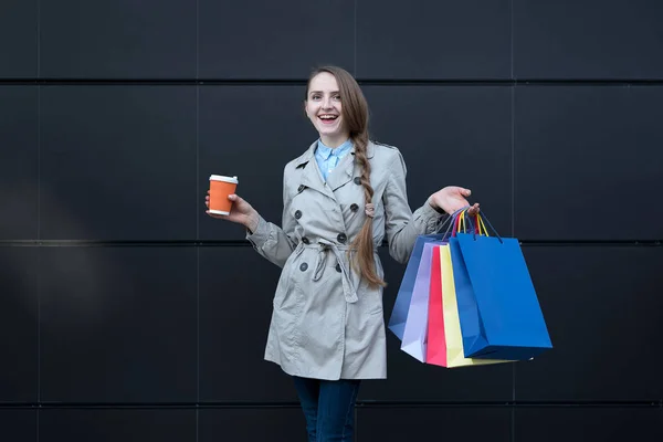 Szczęśliwa młoda kobieta z kolorowymi torbami i papierową filiżanką. Czarna ściana ulicy na tle — Zdjęcie stockowe
