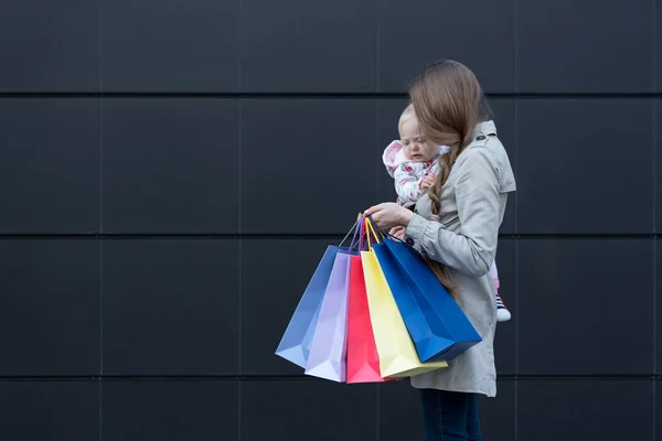 Joven madre con una hija pequeña en los brazos y bolsas de compras en la mano. Negro pared de la calle en el fondo — Foto de Stock