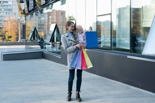 Gelukkige jonge moeder met dochtertje op de armen en boodschappentassen in de hand. Winkeldag. Mall op achtergrond — Stockfoto