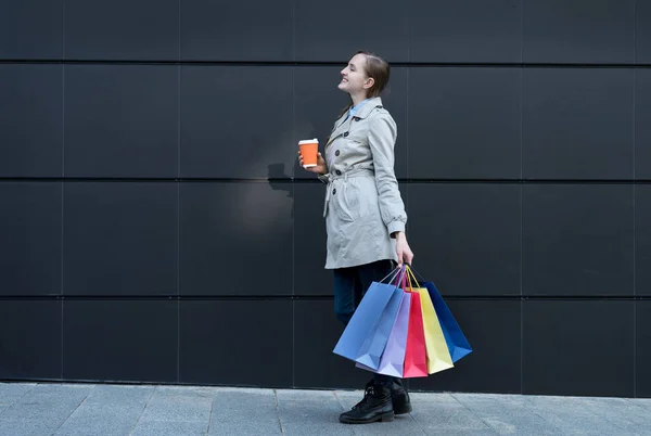 Šťastná mladá žena s barevnými taškami a papírovým kelímkem. Černá ulice zeď na pozadí — Stock fotografie