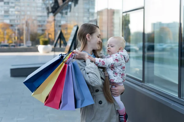 Szczęśliwa młoda matka z córeczką na rękach i torbami na zakupy w ręku. Dzień zakupów. Centrum handlowe na tle — Zdjęcie stockowe