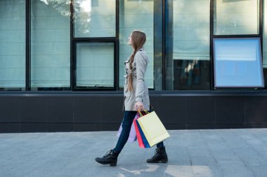 Renkli çantalı genç bir kadın sokakta yürüyor. Arkaplanda alışveriş merkezi
