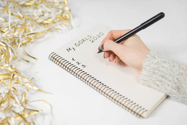 Female hands writing My Goals 2020 in a notebook. Tinsel, New Years concept