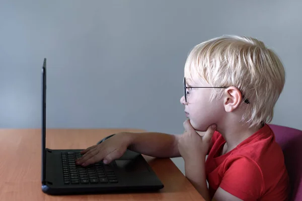 Το αστείο ξανθό αγόρι με τα γυαλιά κάθεται σε ένα λάπτοπ. Ίντερνετ και νηπιαγωγείο — Φωτογραφία Αρχείου