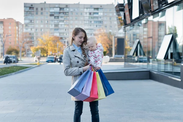 Šťastná mladá matka s malou dcerou na ramenou a nákupními taškami v ruce. Nákupní den. Nákupní centrum na pozadí — Stock fotografie