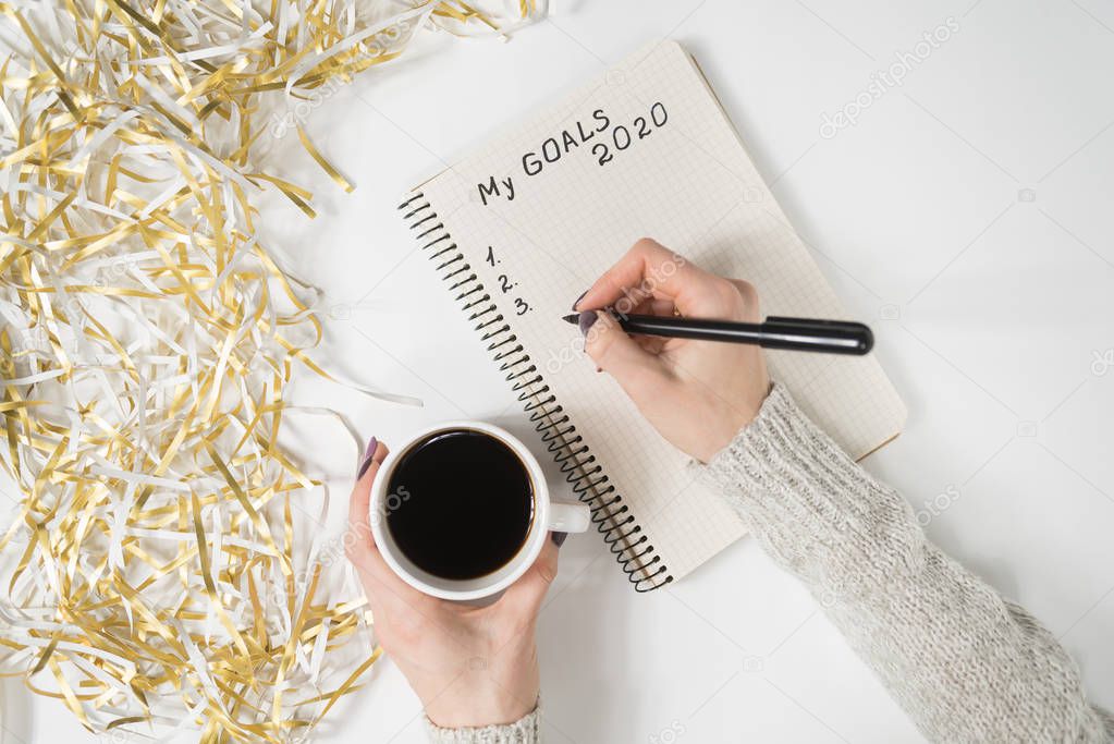 Female hands writing My Goals 2020 in a notebook. Mug of coffee on the table, top view