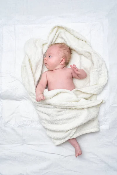 Aktiv baby utan kläder i en vit filt på ett vitt lakan. — Stockfoto
