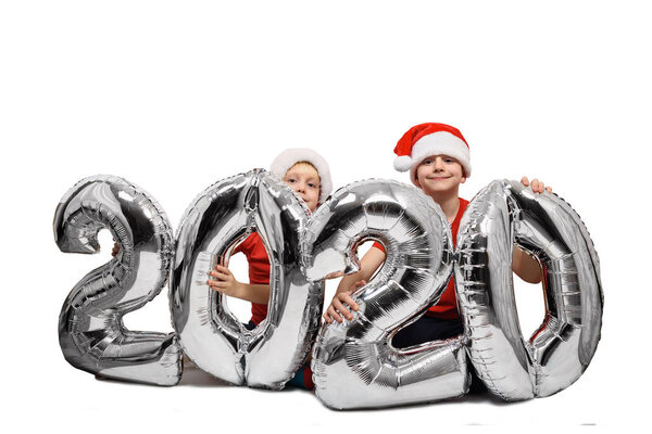Два мальчика в рождественских шляпах держат серебряную надувную фигуру 2020 года. Белый фон. Новогодняя концепция
