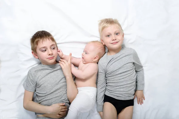 Dos hermanos mayores juegan y se ríen con el bebé más joven en una cama blanca. Feliz infancia, gran familia — Foto de Stock