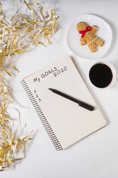 Phrase My Goals 2020 in a notebook, pen. Mug of coffee, gingerbread man and tinsel. Top view