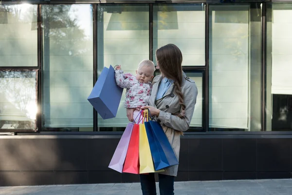 Giovane madre felice con figlioletta sulle braccia e borse della spesa in mano. Giornata dello shopping. Centro commerciale sullo sfondo — Foto Stock