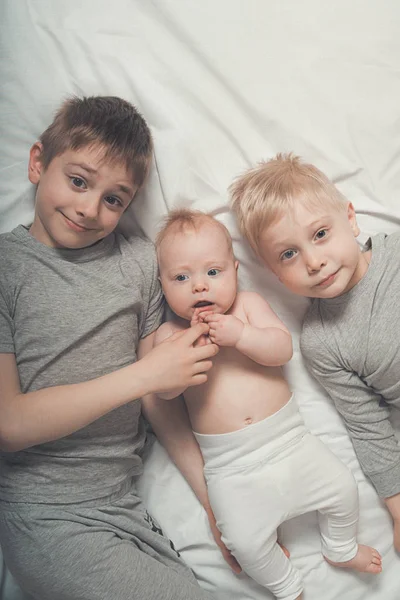 Two older brothers are lying with the youngest baby in a white bed. Happy childhood, big family