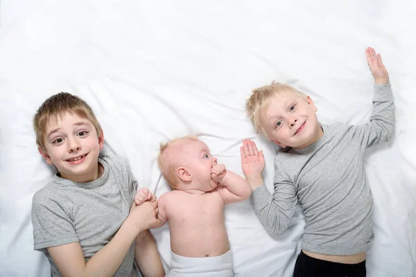 Dos hermanos mayores están acostados con el bebé más joven en una cama blanca. Feliz infancia, gran familia — Foto de Stock