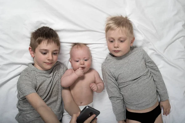 Dos hermanos con un hijo menor están acostados en una cama blanca y tomando selfie en un teléfono inteligente. Feliz infancia, gran familia — Foto de Stock