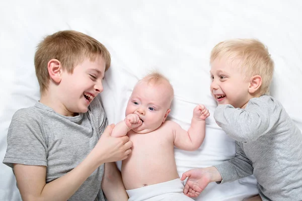 Dos hermanos mayores juegan y se ríen con el bebé más joven en una cama blanca. Feliz infancia, gran familia — Foto de Stock