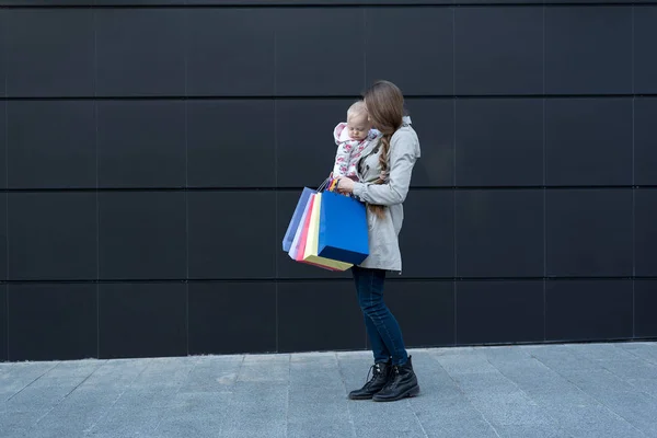 Joven madre con una hija pequeña en los brazos y bolsas de compras en la mano. Negro pared de la calle en el fondo —  Fotos de Stock